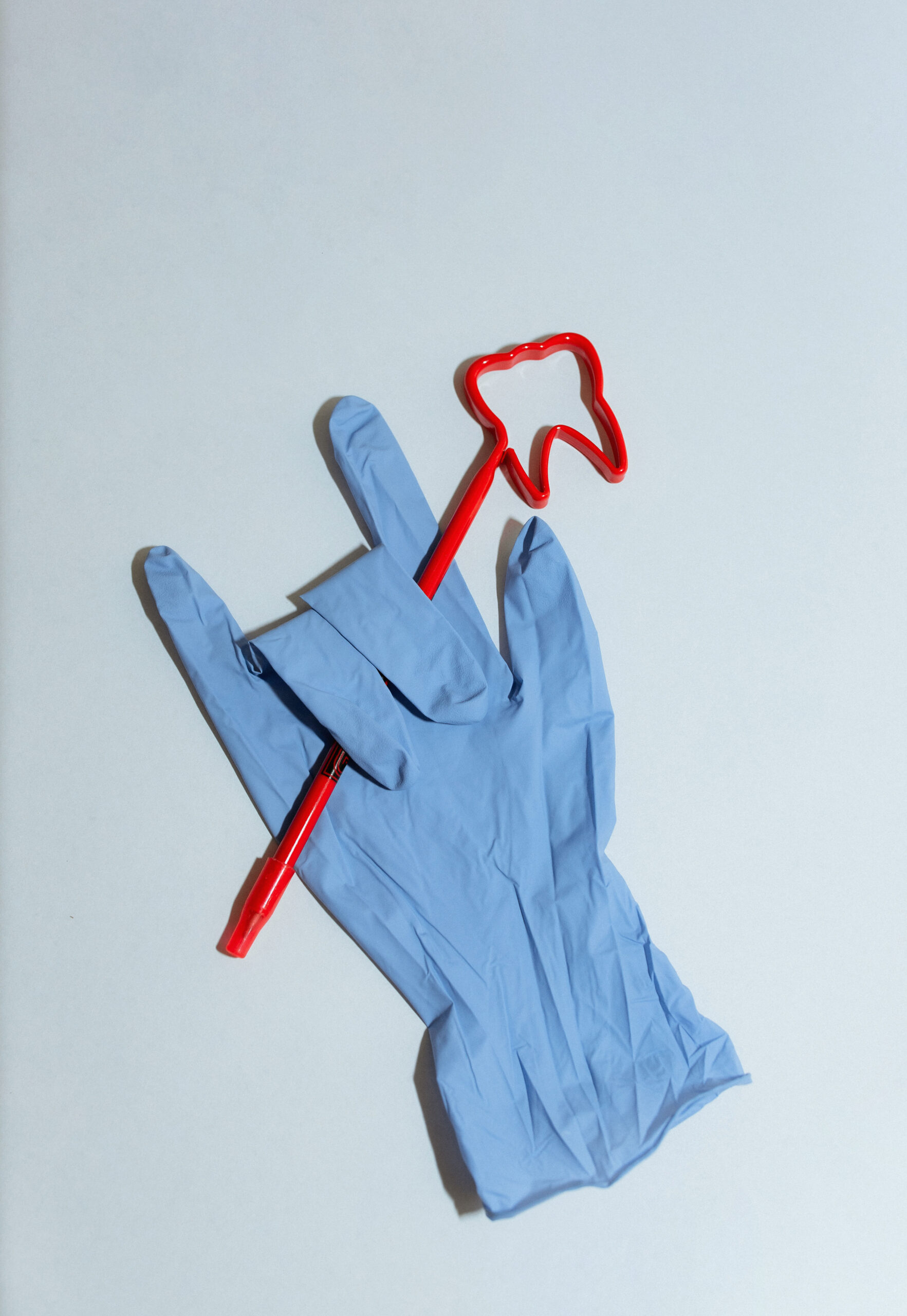 Blue dental exam glove holding a red pen with a tooth-shaped topper, photographed on a white surface